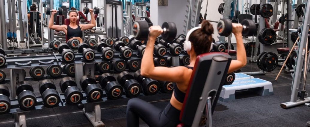 Woman exercising in the gym lifting dumbbells with headphones on for a strength-building workout.